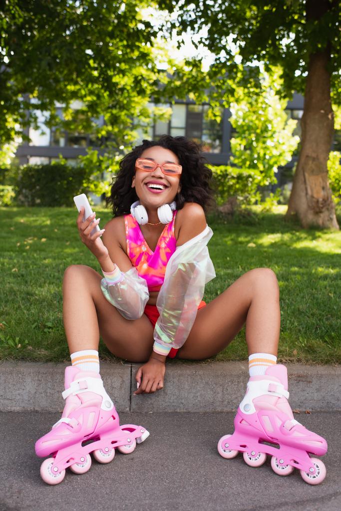щаслива афроамериканка в навушниках і на роликах посміхається, тримаючи смартфон
  - Фото, зображення