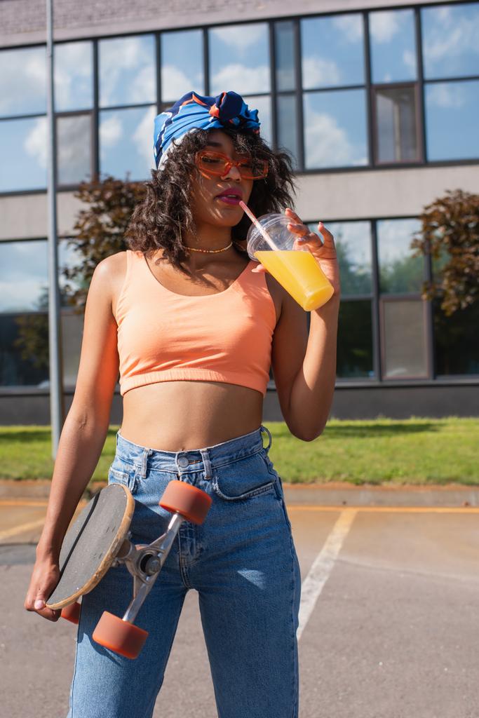 jeune femme afro-américaine en foulard boire du jus d'orange et tenir longboard à l'extérieur  - Photo, image