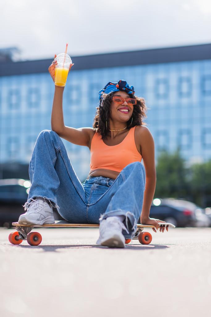 donna afro-americana sorridente in occhiali da sole seduta sul longboard mentre tiene smartphone e tazza di plastica  - Foto, immagini
