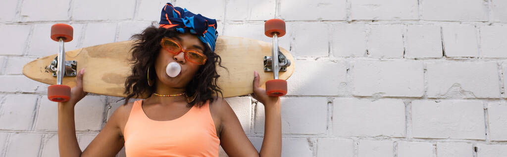 mujer afroamericana con estilo en gafas de sol y pañuelo para la cabeza que sopla goma de mascar y la celebración de longboard cerca de la pared de ladrillo, bandera - Foto, Imagen