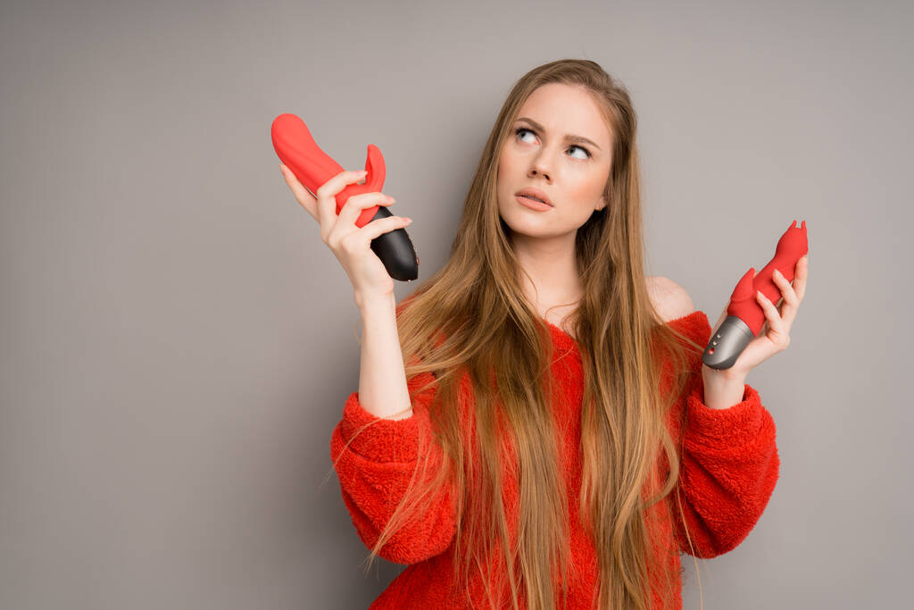 A beautiful woman with long hair chooses a dildo or a vibrator. Two massagers for sex in the hands of the girl. Sex shop concept - Foto, immagini