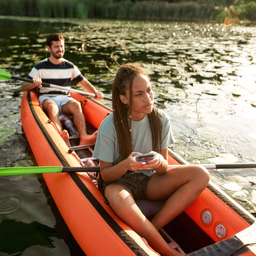 Портрет молодой женщины-путешественницы выглядит смущенно, используя смартфон, проводя время со своим парнем на открытом воздухе, катаясь на байдарках по реке - Фото, изображение