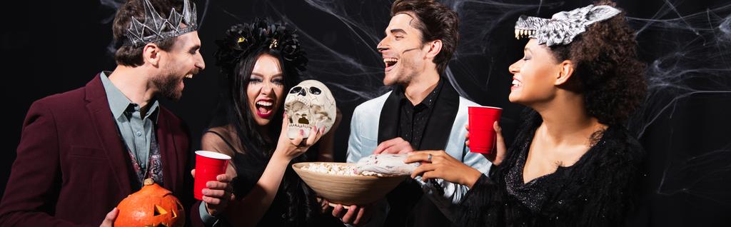 eccitati amici multiculturali bere birra e mangiare popcorn durante la festa di Halloween sul nero, banner - Foto, immagini