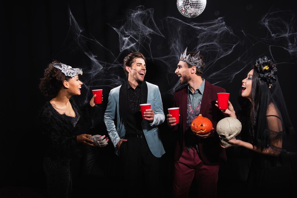 heureux amis multiethniques avec des tasses en plastique parler pendant la fête d'Halloween sur noir  - Photo, image