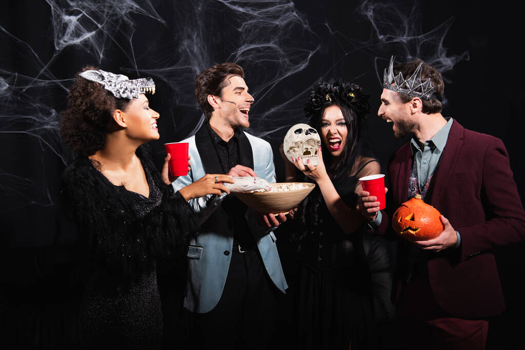 щасливий мультикультурний друг з попкорном і пивом сміється під час вечірки на Хеллоуїн на чорному
 - Фото, зображення