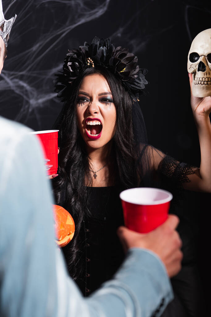 aziatische vrouw in vampier kostuum grommen terwijl angstaanjagende wazig man met griezelige schedel op zwart - Foto, afbeelding
