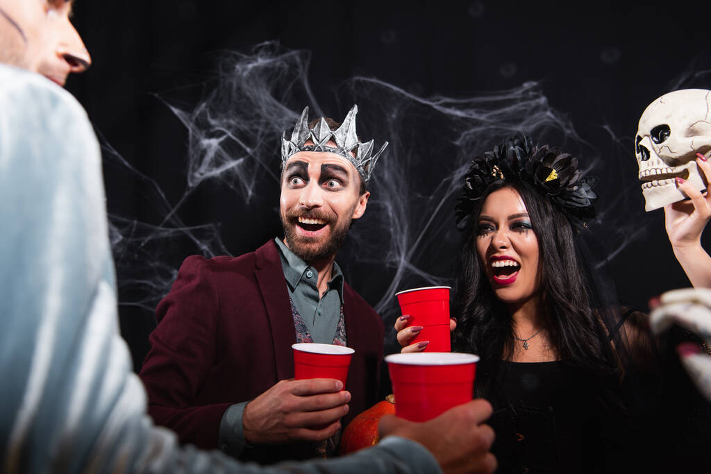 opgewonden interraciale vrienden in halloween kostuums met plastic bekers terwijl het praten op zwart - Foto, afbeelding