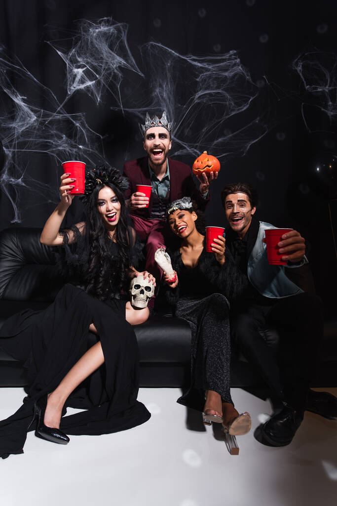 счастливые многонациональные друзья в костюмах на Хэллоуин, смотрящие в камеру, сидя на диване на черном - Фото, изображение