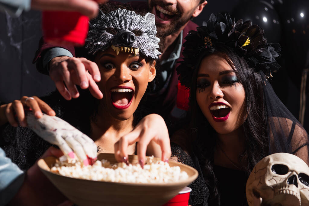 excited interracial women in halloween costumes, with skull and toy hand near bowl with popcorn - Photo, Image