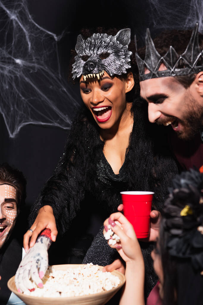 Веселая африканская американка, принимающая попкорн с игрушечной рукой рядом с мужчиной в костюме вампира на Хэллоуин на черном - Фото, изображение