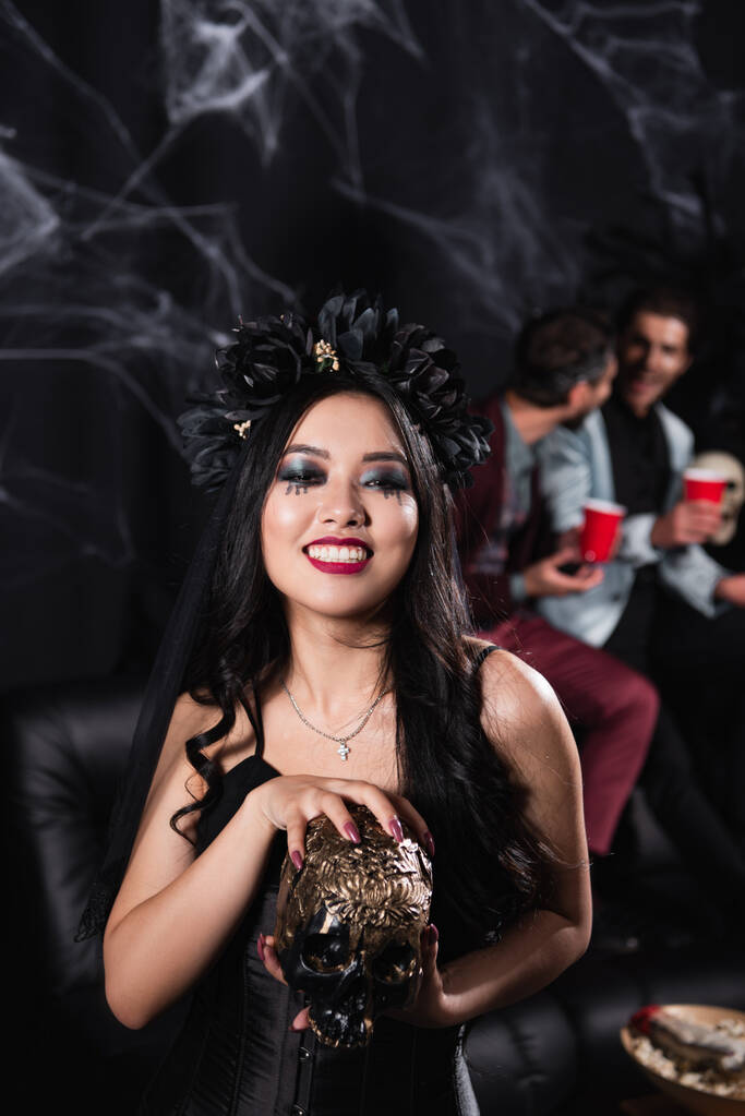 smiling asian woman in vampire halloween costume holding spooky skull on black - Photo, Image