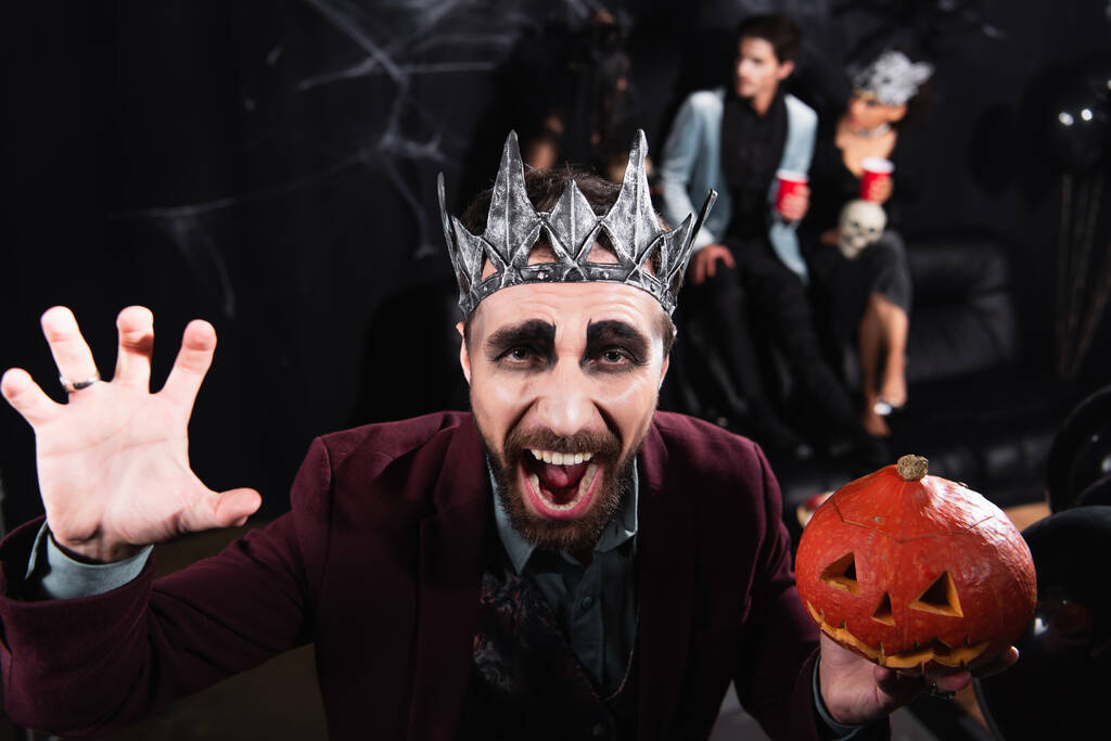 Mann in Vampir-Königskrone hält geschnitzten Kürbis in der Hand und knurrt während der Halloween-Party auf schwarz in die Kamera - Foto, Bild