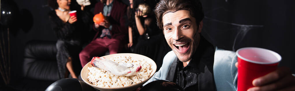 веселий чоловік з макіяжем на Хеллоуїн тримає миску з попкорном та іграшковою рукою біля розмитих друзів на чорному фоні, банер
 - Фото, зображення