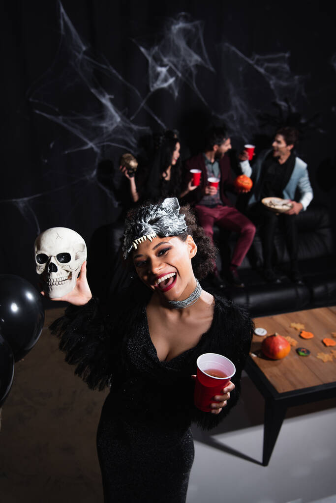cheerful african american woman in wolf mask holding spooky skull while looking at camera on black - Photo, Image