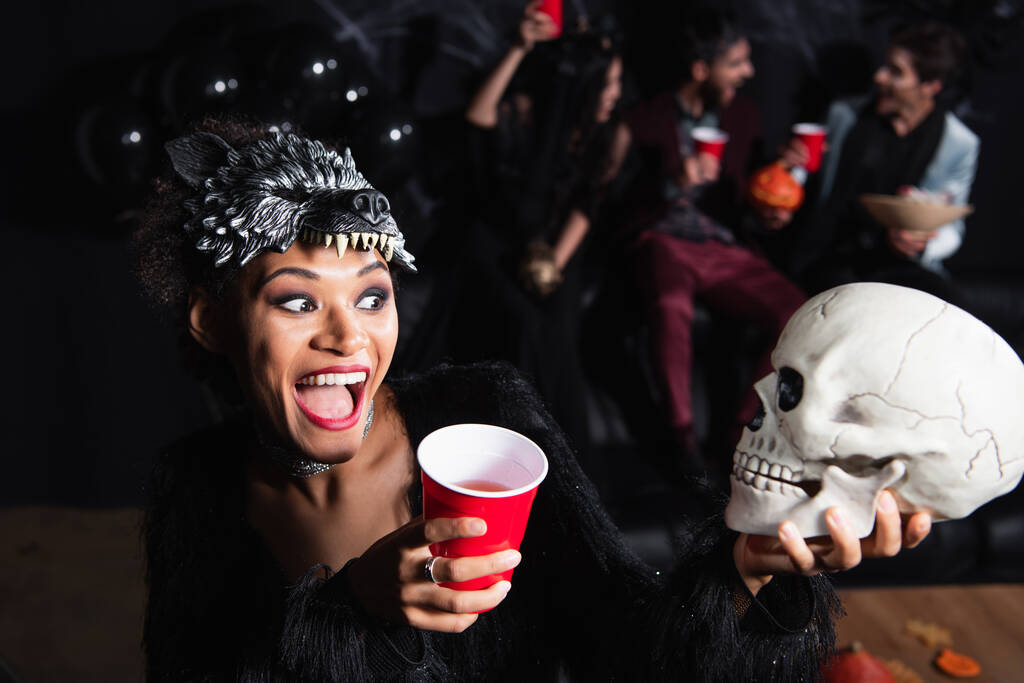 amazed african american woman in wolf mask looking at scary skull near blurred friends on black background - Photo, Image
