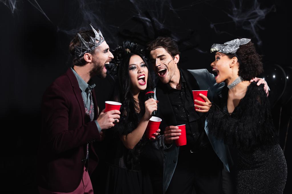 excited multiethnic friends in halloween costumes singing karaoke on black   - Photo, Image