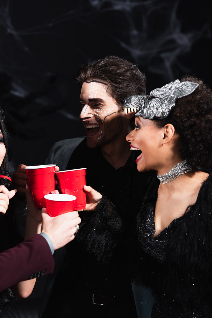 excité afro-américaine femme cliquetis tasses en plastique avec des amis chantant karaoké sur fête d'Halloween sur noir  - Photo, image