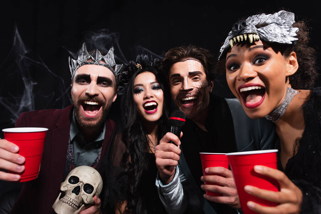 podekscytowani wielonarodowymi przyjaciółmi patrzącymi w kamerę podczas śpiewania karaoke na halloween party na czarno  - Zdjęcie, obraz