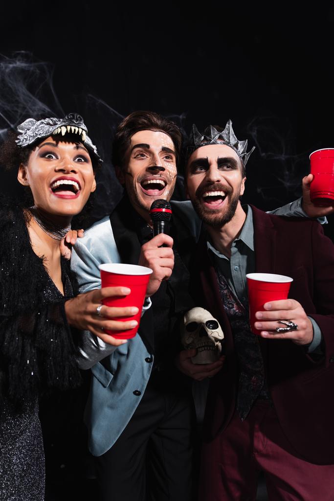 amigos multiétnicos felizes em trajes de Halloween cantando karaoke no preto - Foto, Imagem