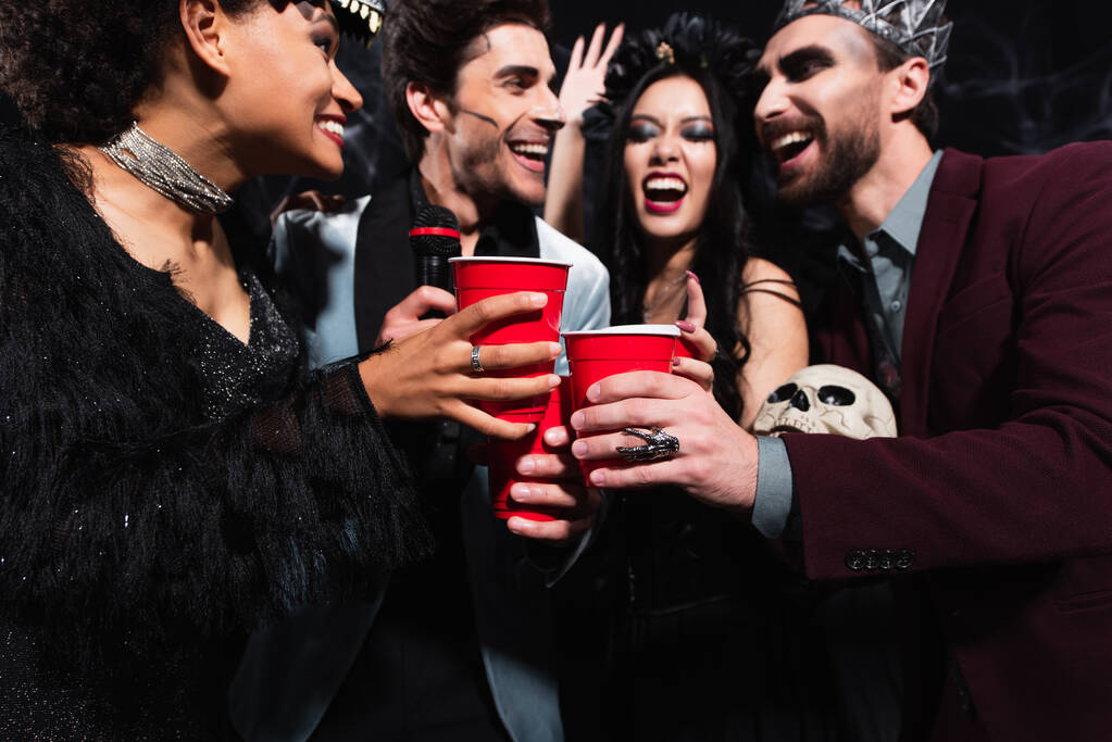 alegres amigos multiétnicos brindando con copas de plástico mientras cantan karaoke en negro  - Foto, Imagen