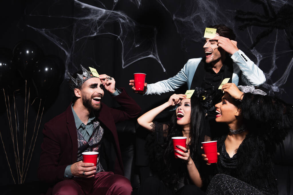 hombre excitado señalando a amigo en la corona del rey vampiro jugando adivina quién juego durante la fiesta de Halloween en negro - Foto, imagen