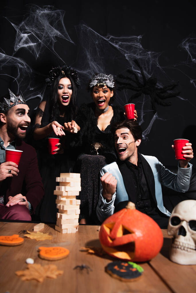 aufgeregter Mann in Halloween-Make-up zeigt Win-Geste in der Nähe von Holzklötzen-Spiel und multiethnischen Freunden auf Schwarz   - Foto, Bild