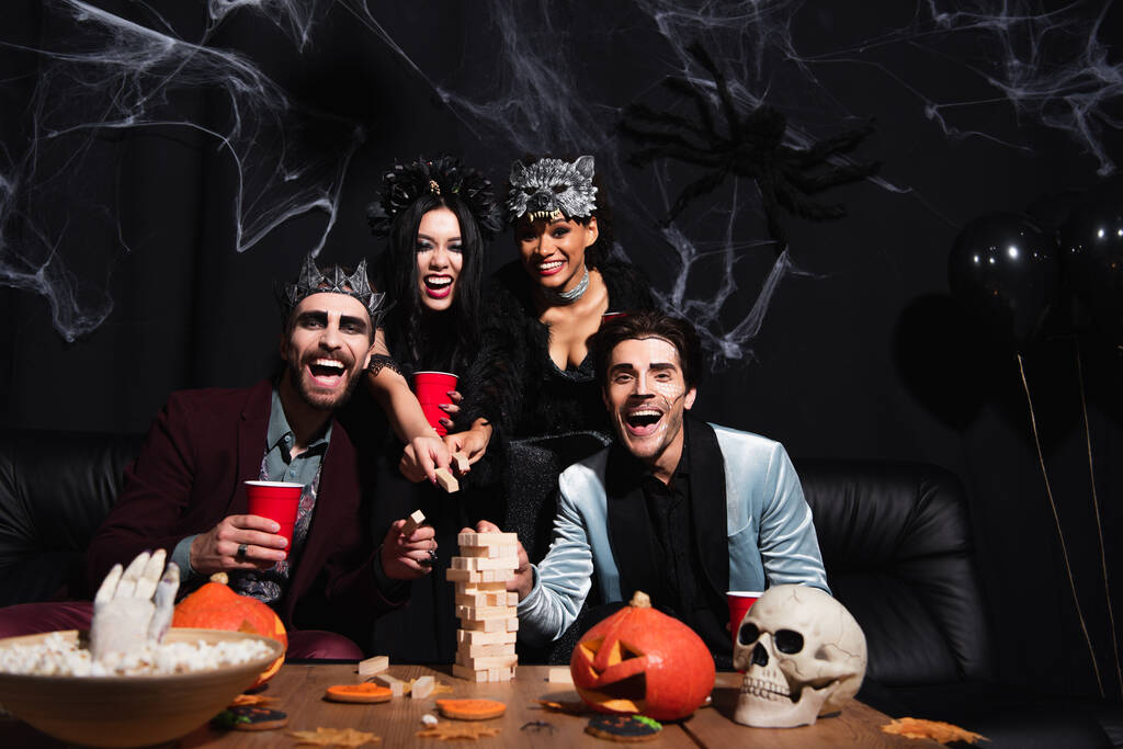 eccitate donne multiculturali in costumi di Halloween giocare blocchi di legno gioco con gli uomini su nero  - Foto, immagini