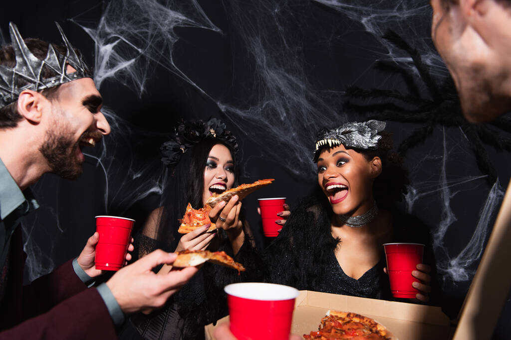изумленная африканская американка, держащая пиццу рядом с многонациональными друзьями в костюмах на Хэллоуин на черном - Фото, изображение