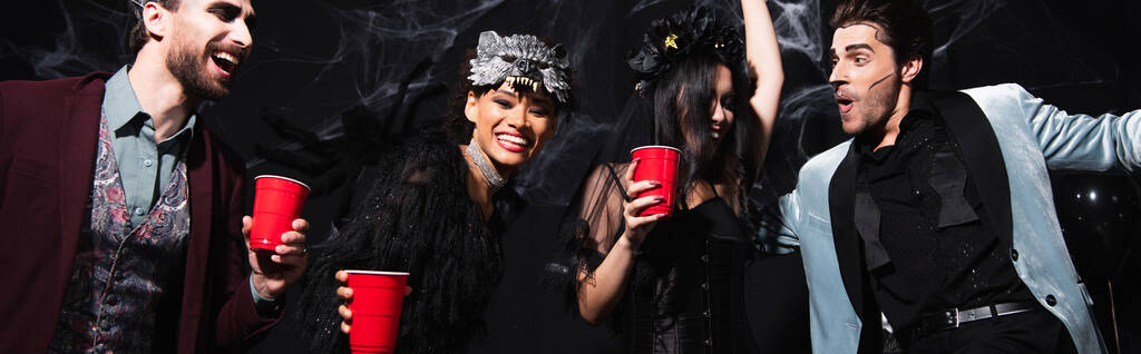 fröhliche multikulturelle Freunde in Halloween-Kostümen tanzen bei Party auf schwarzem Banner - Foto, Bild