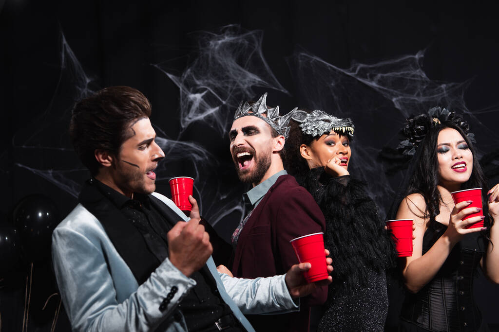 vidám multietnikus barátok műanyag poharak táncolnak halloween party a fekete   - Fotó, kép