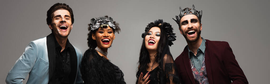 alegres amigos multiétnicos en disfraces de Halloween riendo de cámara aislada en gris, pancarta - Foto, imagen