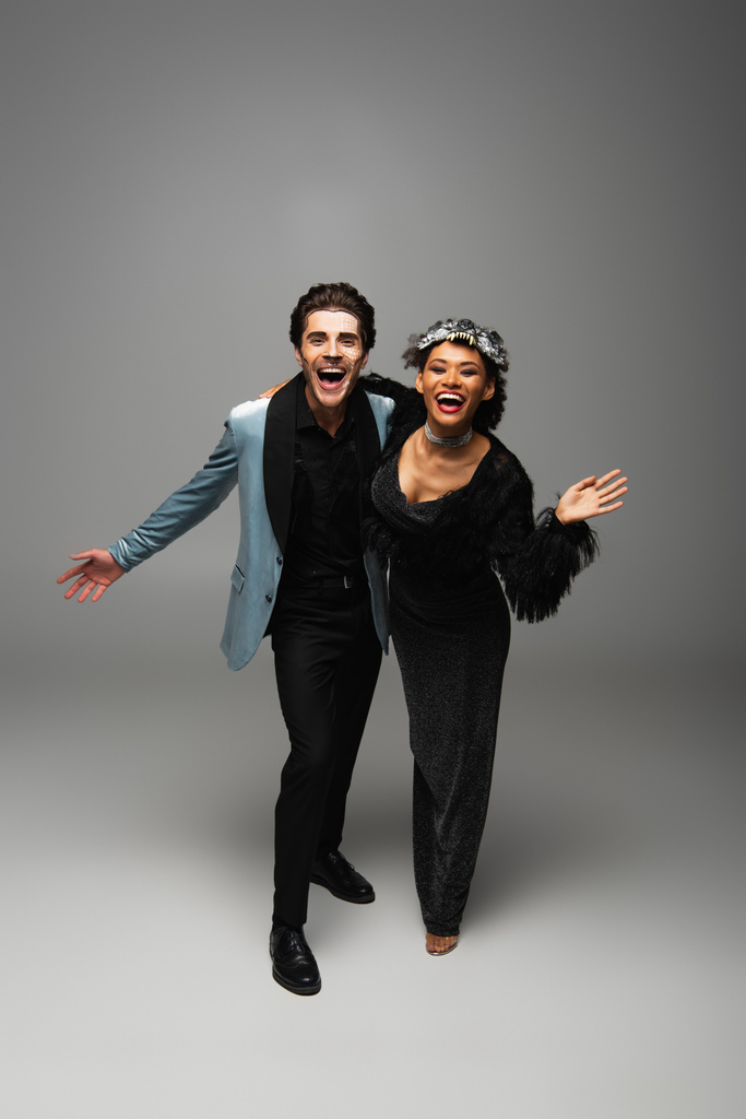 excited african american woman in werewolf mask waving hand near laughing man on grey - Photo, Image