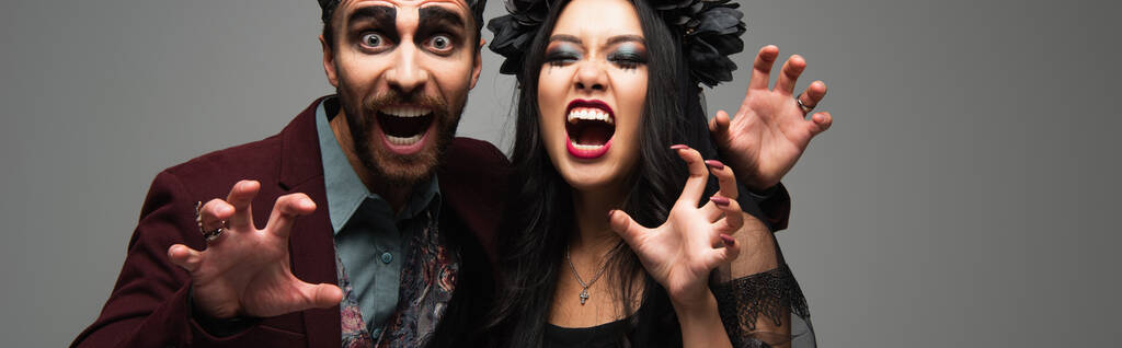 straszne międzyrasowe para w halloween kostiumy pokazujące straszne gesty i warczenie izolowane na szary, sztandar - Zdjęcie, obraz