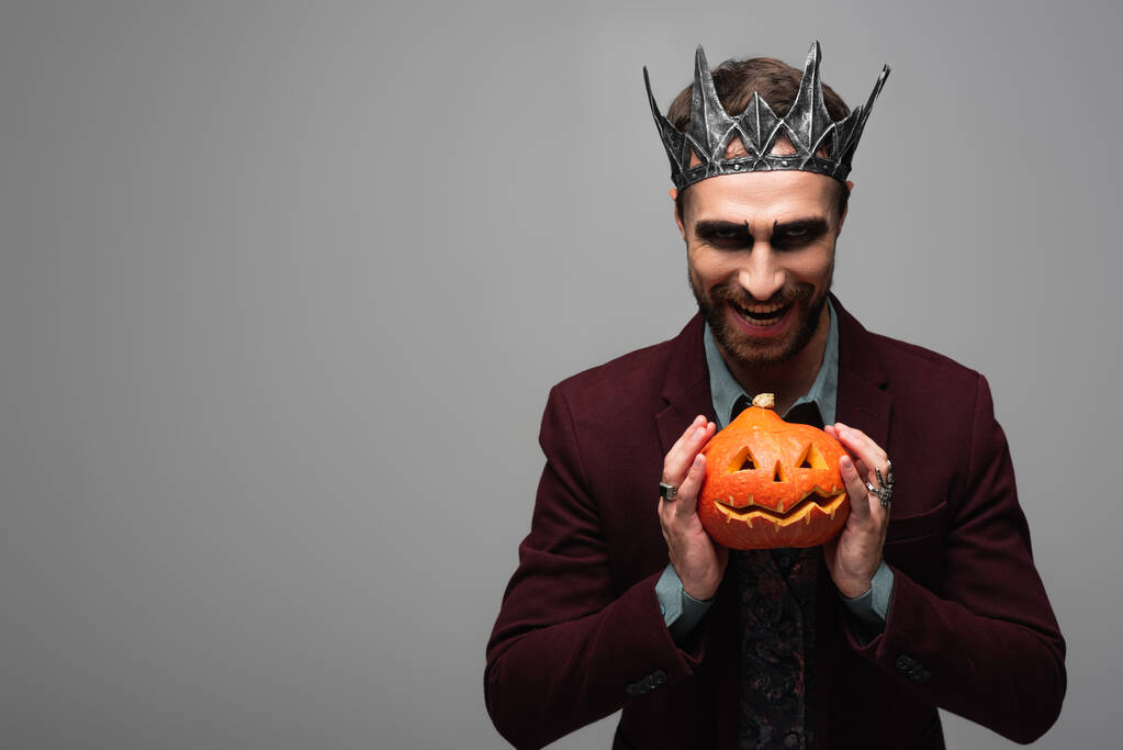 саркастичный человек в короне вампира на Хэллоуин улыбается и показывает жуткую тыкву, изолированную на серой - Фото, изображение