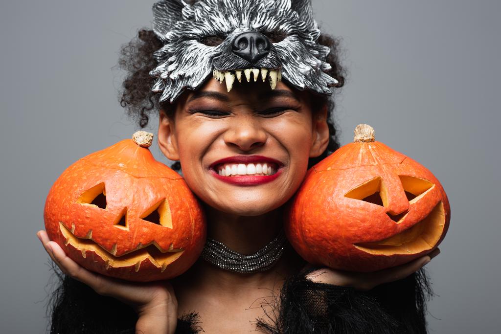 overjoyed Afryki amerykanka kobieta w wilk halloween maska gospodarstwa rzeźbione dynie i uśmiech z zamkniętymi oczami izolowane na szary - Zdjęcie, obraz