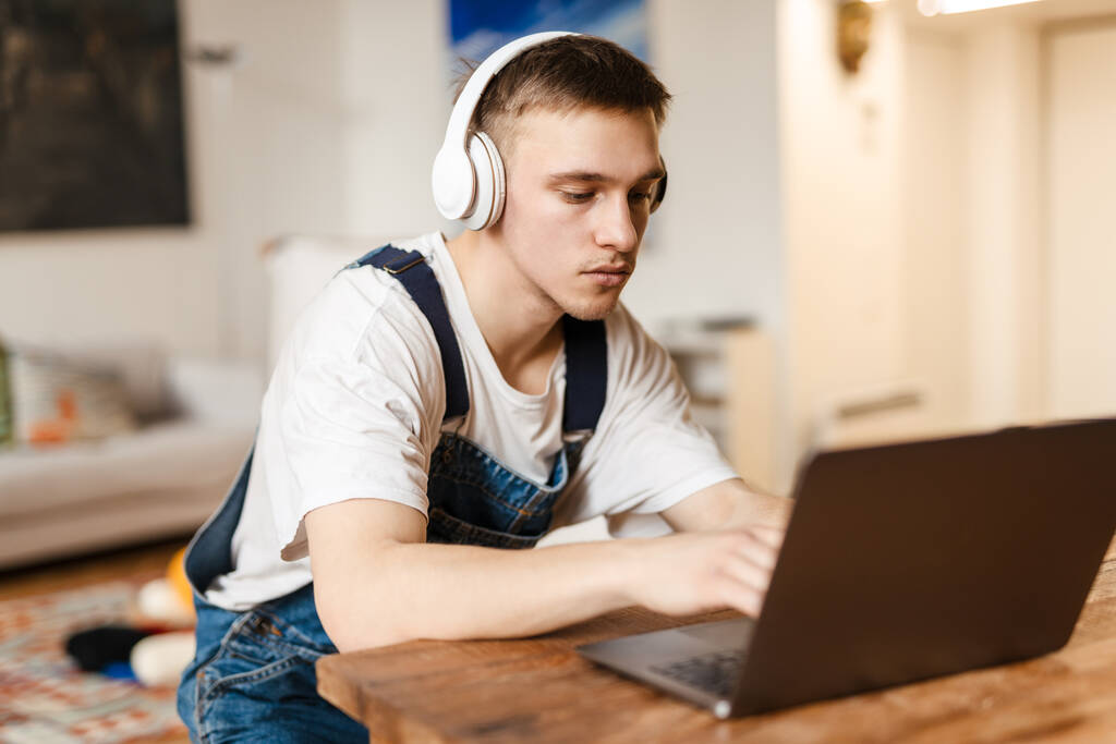 Giovane uomo in cuffia utilizzando il computer portatile mentre si lavora sul tappeto artigianale a casa - Foto, immagini