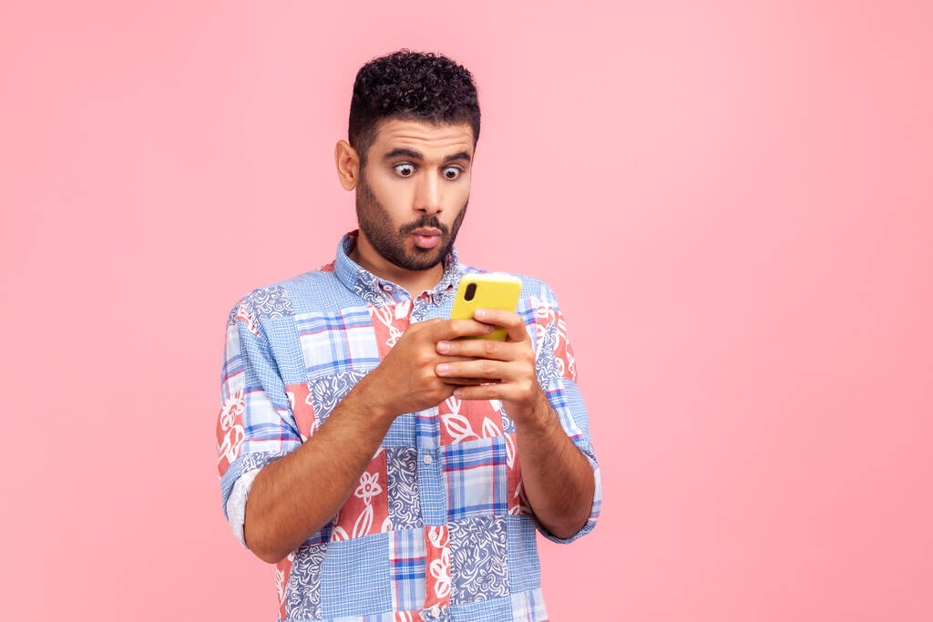 Mobile connection. Portrait of shocked astonished man in blue shirt reading post on social network using cell phone, chatting looking surprised. Indoor studio shot isolated on pink background. - Photo, Image