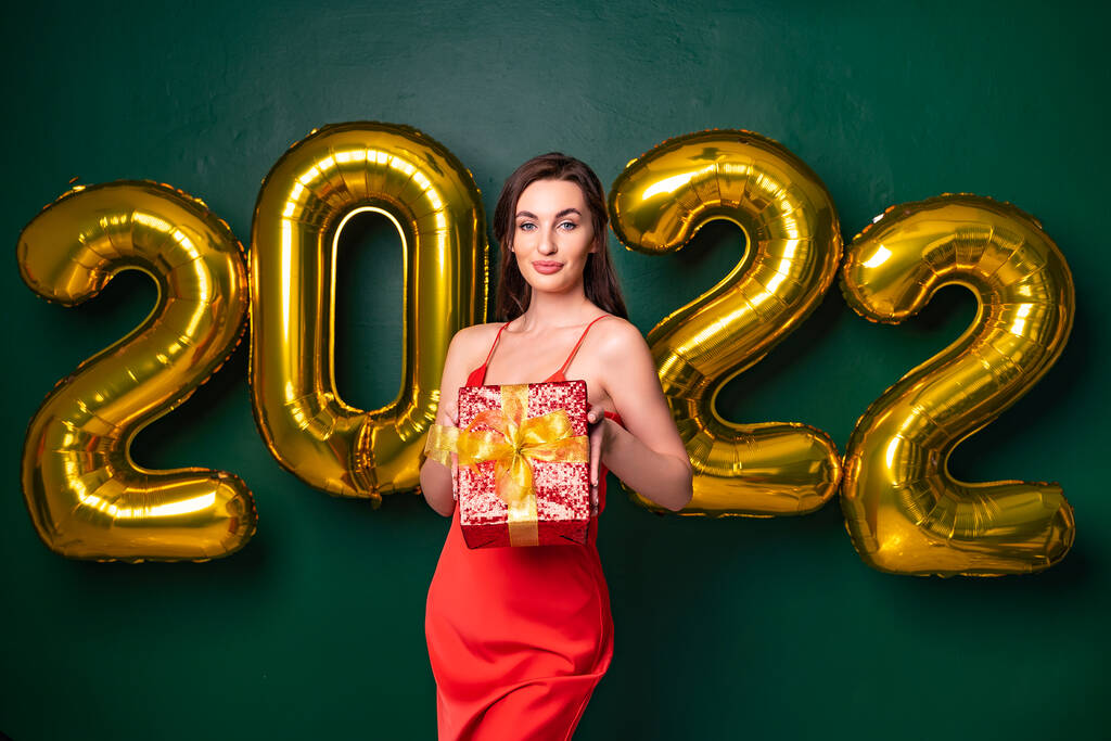 Lächelnde brünette Frau im roten Kleid hebt die rote Geschenkbox in die Höhe. NEUJAHR 2022 goldene Luftballons.  - Foto, Bild
