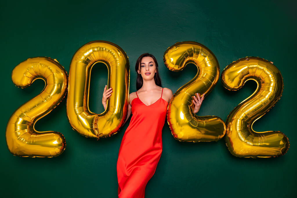 緑の背景に2022年の黄金の風船を保持赤いドレスの若い幸せな女性。新年のコンセプト. - 写真・画像