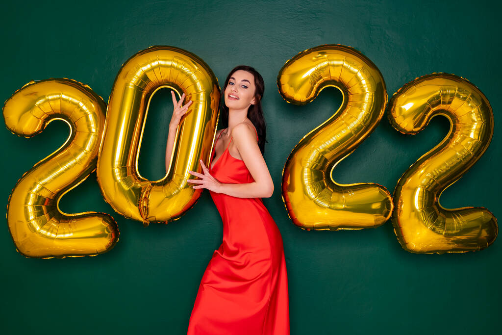 Молодая счастливая женщина в красном платье с 2022 золотыми шариками на зеленом фоне. Концепция Нового года. - Фото, изображение