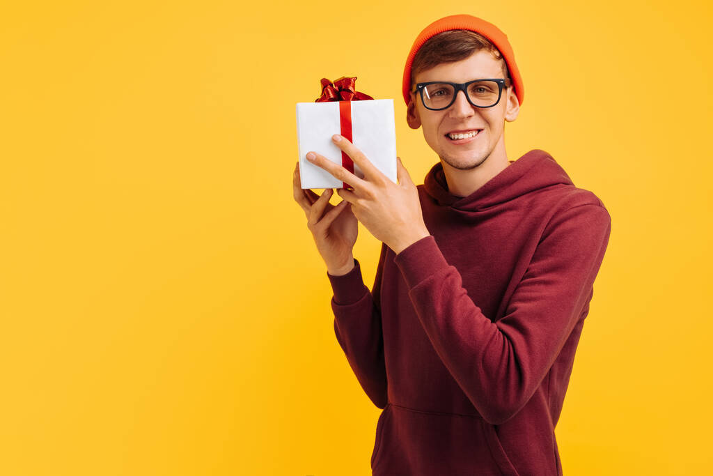 een knappe jongeman met een oranje hoed met bril en een rode trui, denkend aan wat er in zijn cadeau zit, een feestelijke stemming, een nieuwjaarscadeau, handen met een witte kurk en een rode strik - Foto, afbeelding