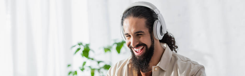 hombre hispano emocionado riendo mientras escucha música en auriculares inalámbricos, pancarta - Foto, Imagen