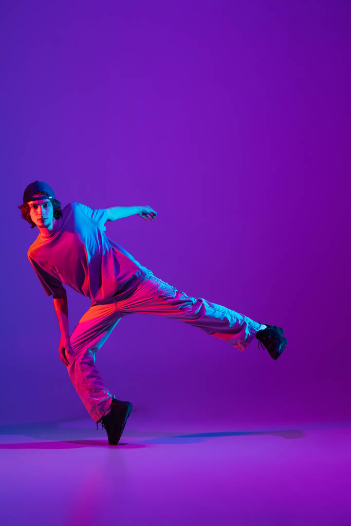 Élégant garçon sportif dansant hip-hop dans des vêtements élégants sur fond coloré à la salle de danse dans la lumière au néon. Culture, mouvement, style et mode des jeunes, action. - Photo, image