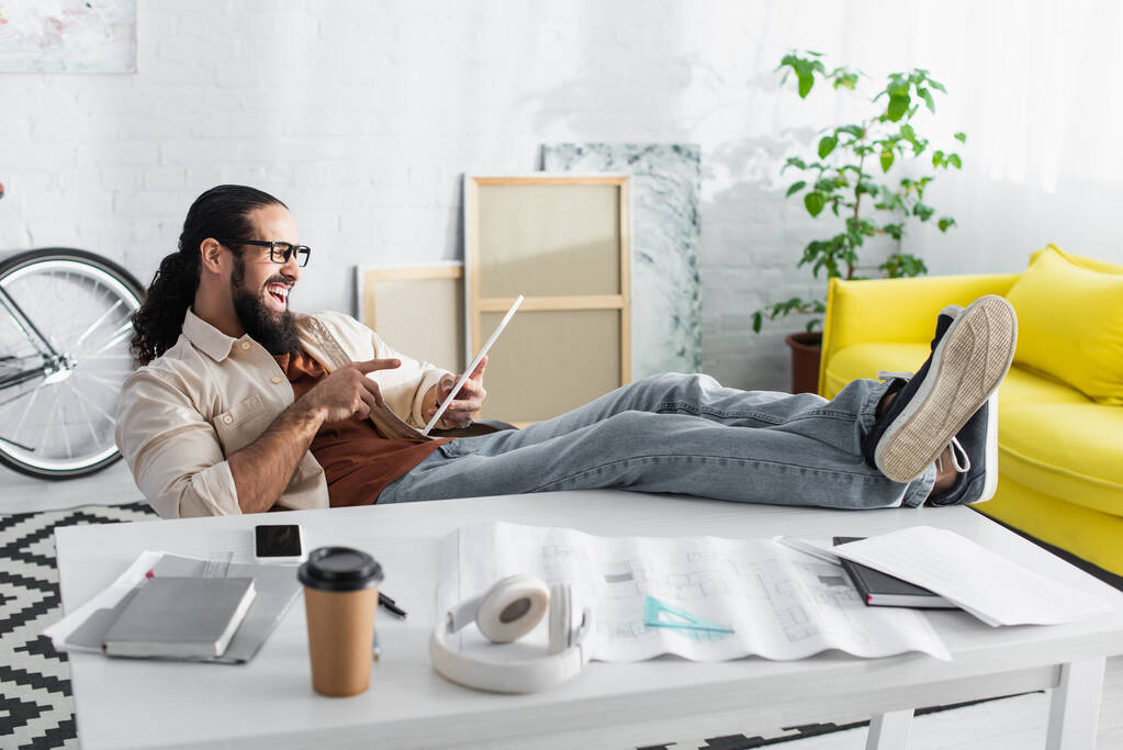 γελώντας Ισπανόφωνος σχεδιαστής δείχνει ψηφιακή ταμπλέτα, ενώ κάθεται με τα πόδια στο γραφείο στο σπίτι - Φωτογραφία, εικόνα