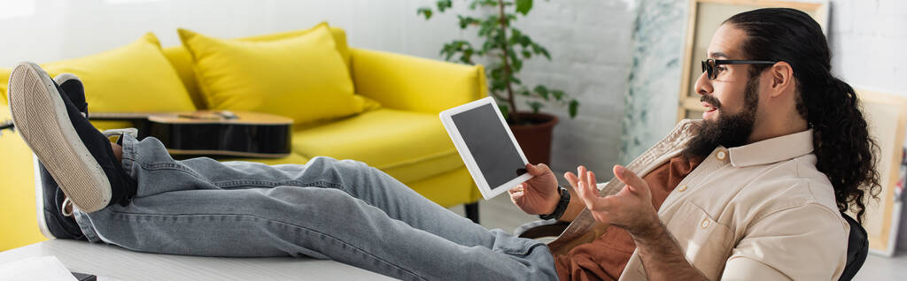 hombre hispano haciendo gestos mientras está sentado con las piernas en el escritorio durante el chat de vídeo en la tableta digital, pancarta - Foto, Imagen