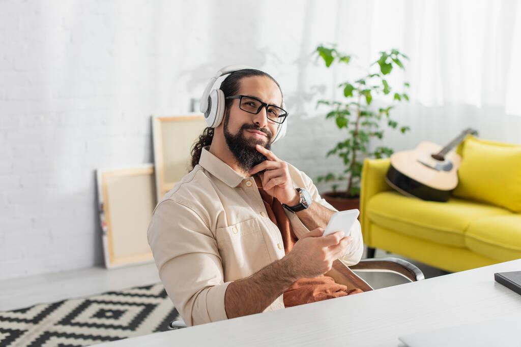 marzycielski i uśmiechnięty Hiszpan ze smartfonem słuchającym muzyki w słuchawkach w domu - Zdjęcie, obraz