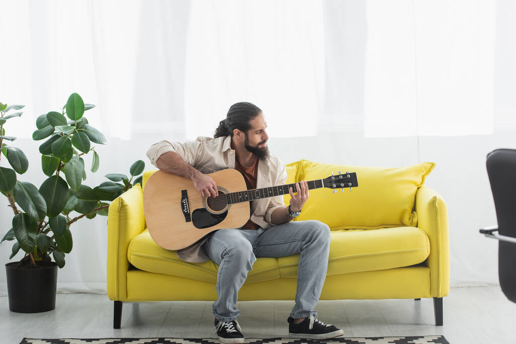 Ansicht des lateinischen Mannes in voller Länge, der auf einer gelben Couch im Wohnzimmer sitzt und Gitarre spielt - Foto, Bild