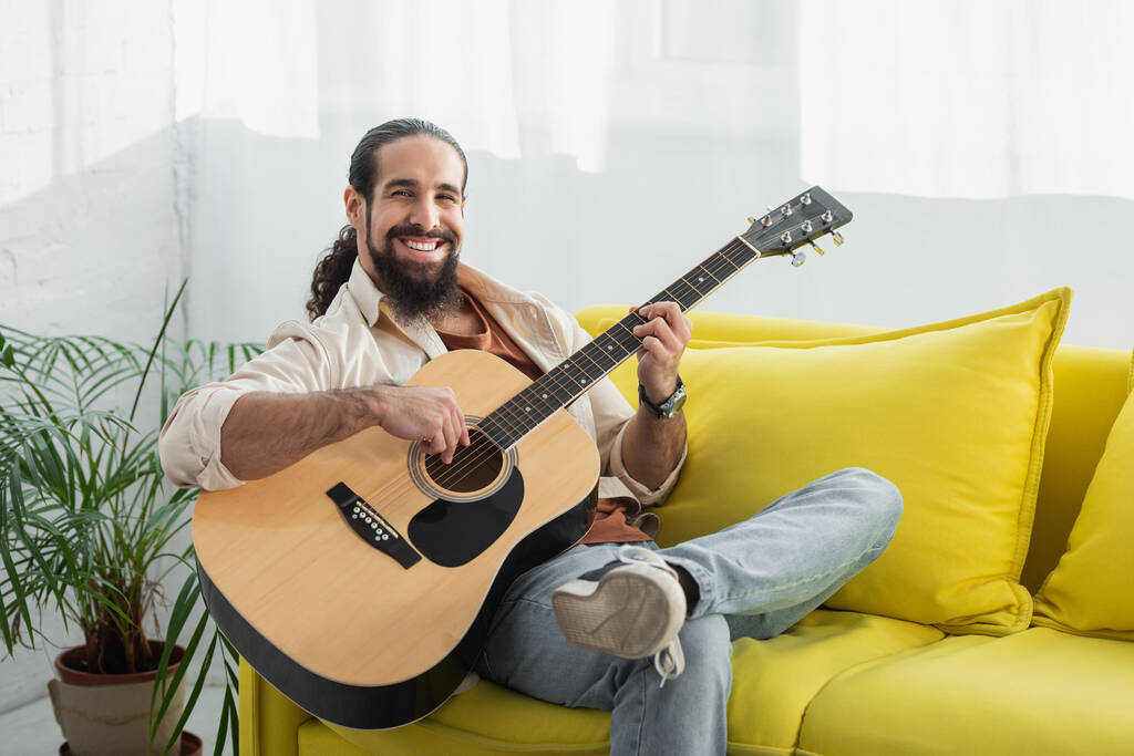 šťastný latin muž při pohledu na kameru při hraní na kytaru na žluté pohovce doma - Fotografie, Obrázek