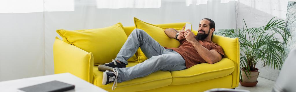 黄色いソファに横になりスマートフォンやバナーでチャットしているラテン系の男性の完全な長距離ビュー - 写真・画像
