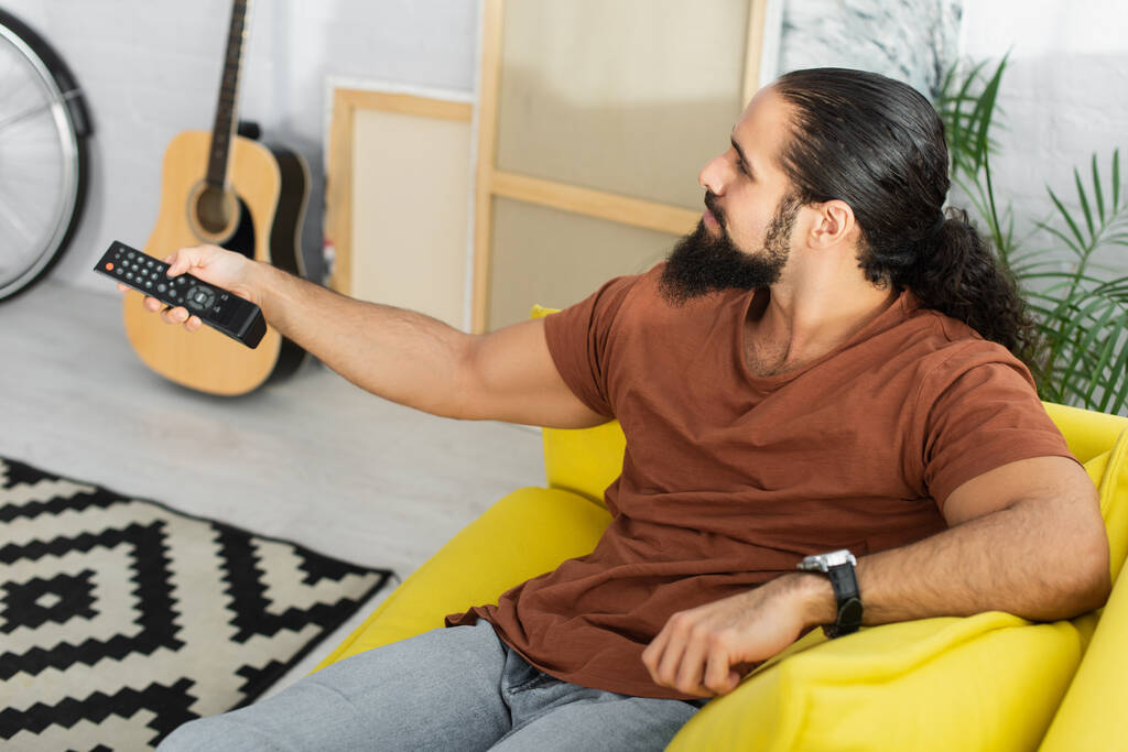 άποψη υψηλής γωνίας του ισπανόφωνου άνδρα κλικ τηλεοπτικά κανάλια κοντά θολή ακουστική κιθάρα και καμβάδες - Φωτογραφία, εικόνα
